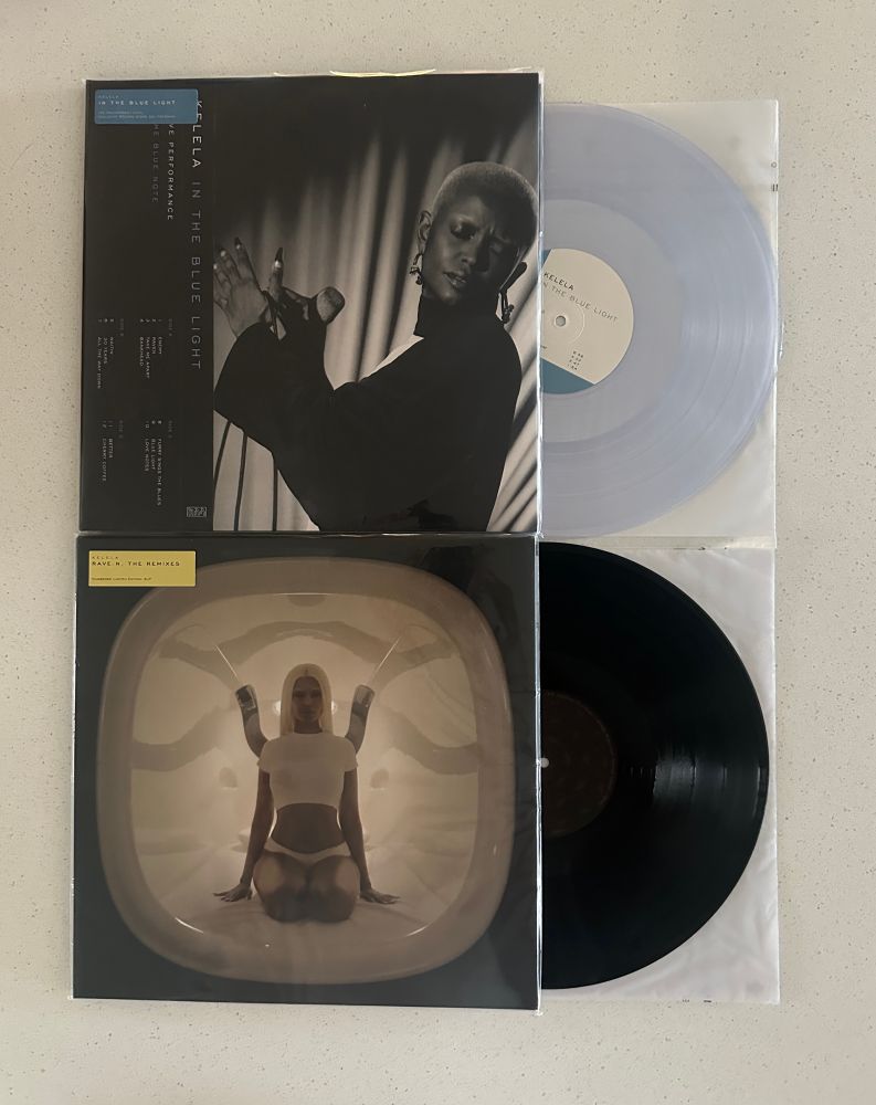 Two vinyl records stacked on top of each other on a white counter, the top a clear pressing of Kelela’s In The Blue Light, the bottom a black pressing of RAVE:N, the remixes by Kelela 