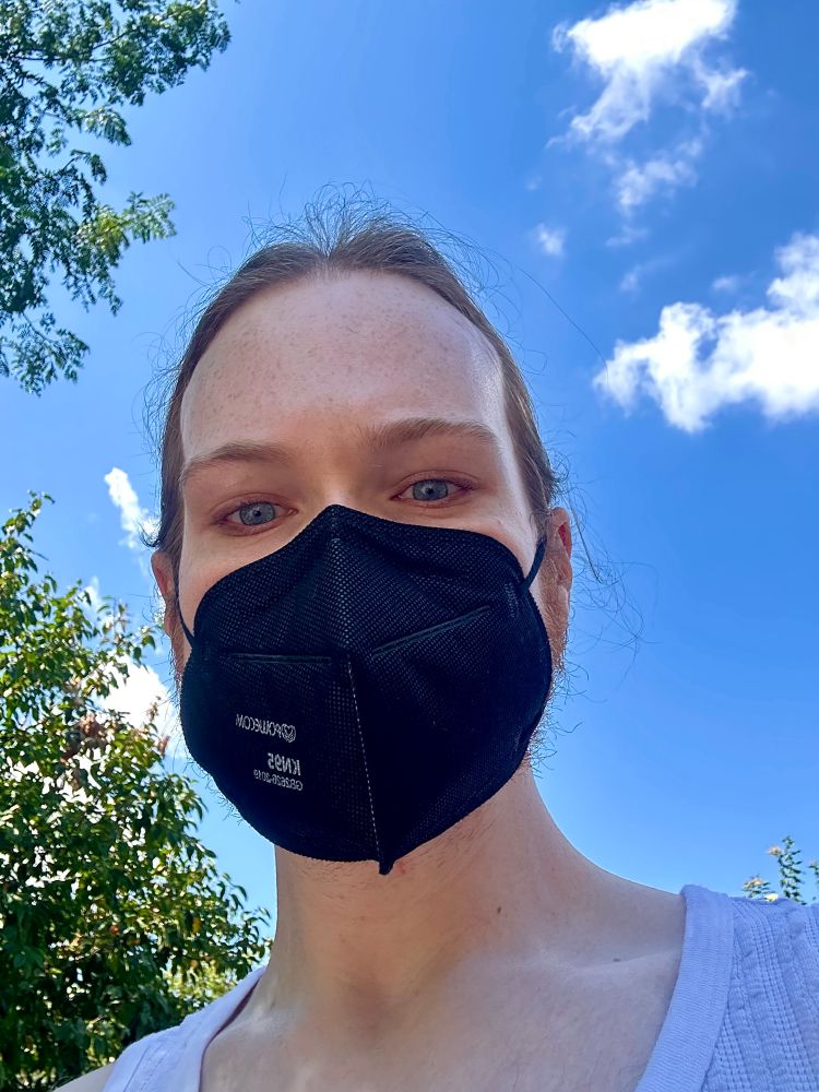picture is a selfie of me, i am wearing a black KN95 mask and my hair is pulled back, you can see i am wearing a white tank top, and the background is trees to my side and the blue sky, with clouds sprinkled around 