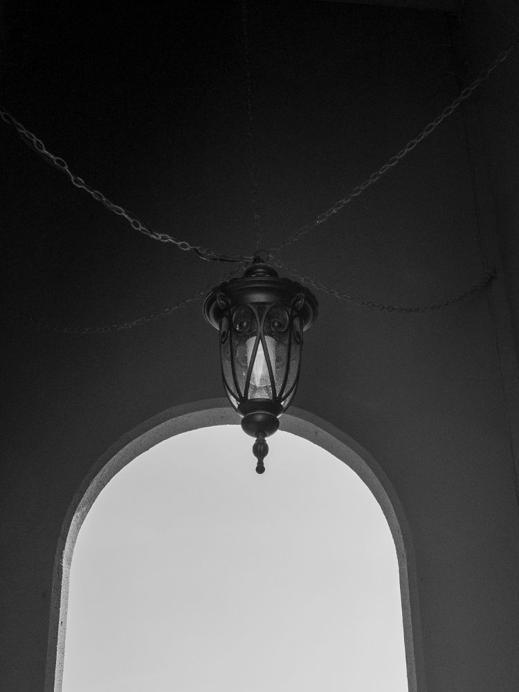 A black and white photograph of a lamp hanging by a chain with four more chains linked to the base , attached to the stone walls. An bright arch is in the background.