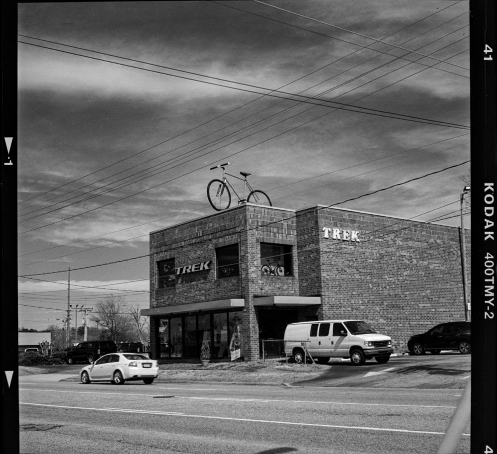 A black and white photograph of a Trek bicycle shop with a large bicycle sitting on top of the roof. A whole bunch of power lines going every way. Dynamic looking sky.