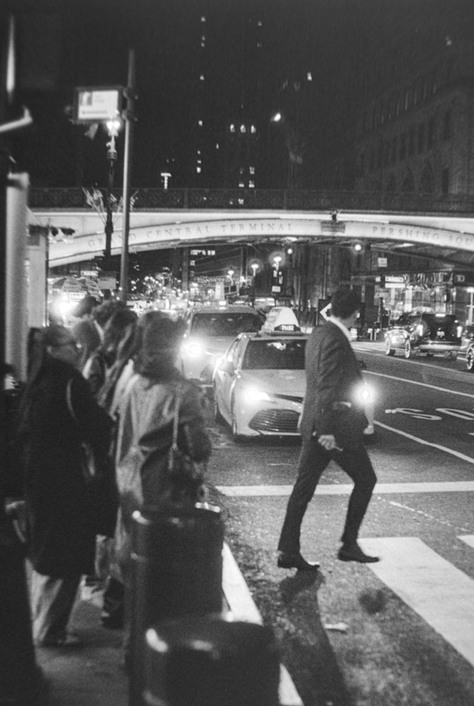 A black and white photograph of a man in a suit crossing the street at a crosswalk at night. Several cars are waiting for him to pass. Other people are standing waiting on the walkway.