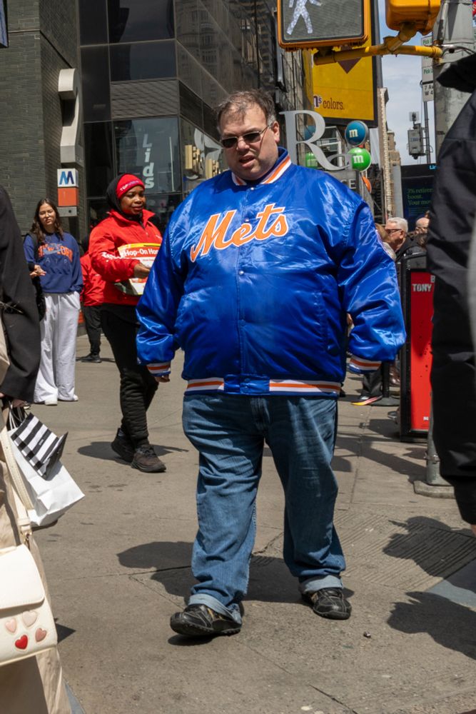A color photograph of a large man with a Mets jacket on walking through the streets of NYC.