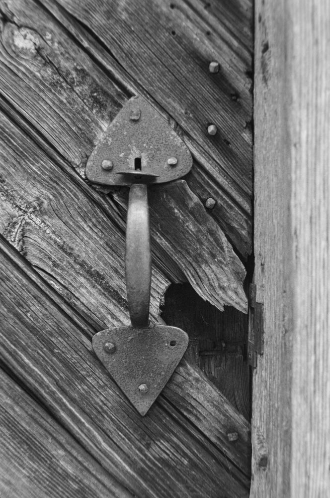 A black and white photograph of a broken old door handle. The wood behind it has been broken away to reviel the absence of a mechanism underneath.