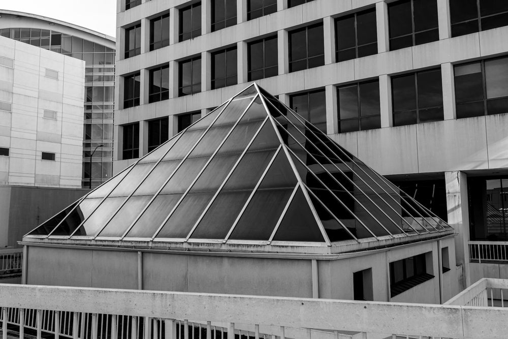 A black and white photograph of a pyramid made of black tented windows in front of a building with more tented windows.