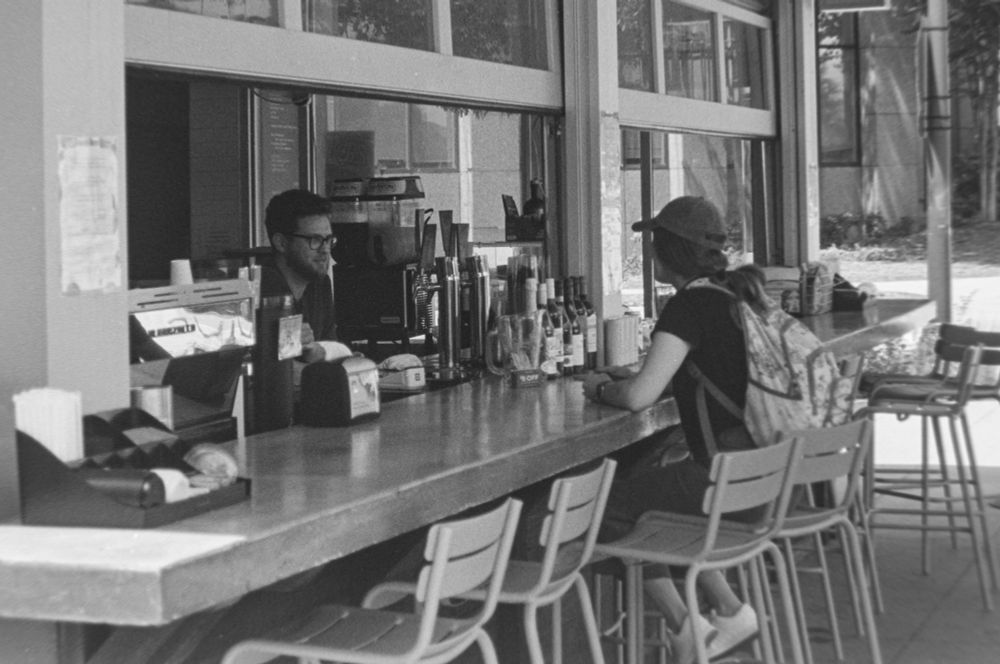 A black and white photograph of a woman in a baseball cap ordering from a guy in glasses at a outdoor coffee bar.