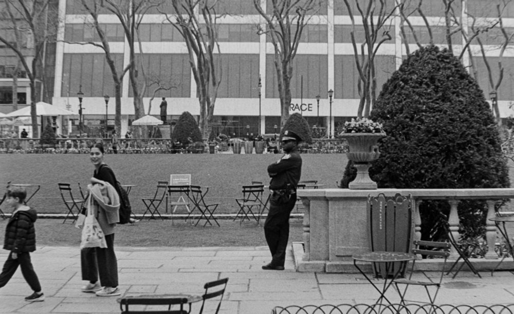 A black and white photograph of a mother and son walking past a police officer in a New York City park. The cop is leaning against a stone rail. Above his head, trees line the background and behind there is a store sign. The trees hid the first letter where all can be read is "RACE".