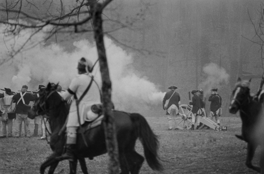 A black and white photograph of a solder on horseback riding by while a canon goes off in the background to the right, and solders are shooting on the left. A thin tree splits the whole photo down the middle.