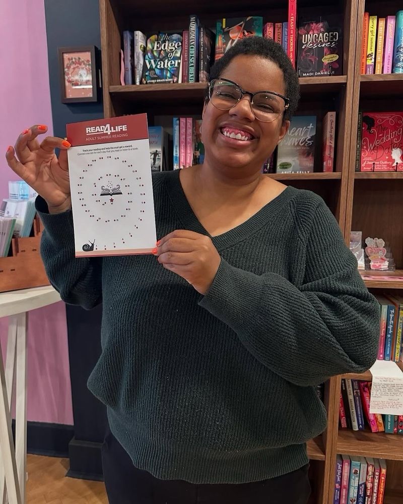 Katherine wearing a dark green sweater holding up a Read 4 Life gameboard in front a bookshelf.