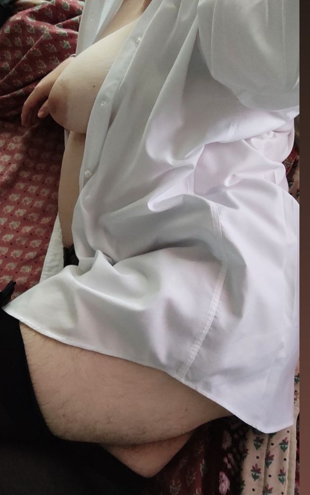 Photo of Darling laying down on bed on their side, leg bent slightly. They're only wearing a white shirt, black underwear and black thigh-high stockings. The shirt is open and their tits and belly are fully exposed, as well as their thick, hairy thighs.