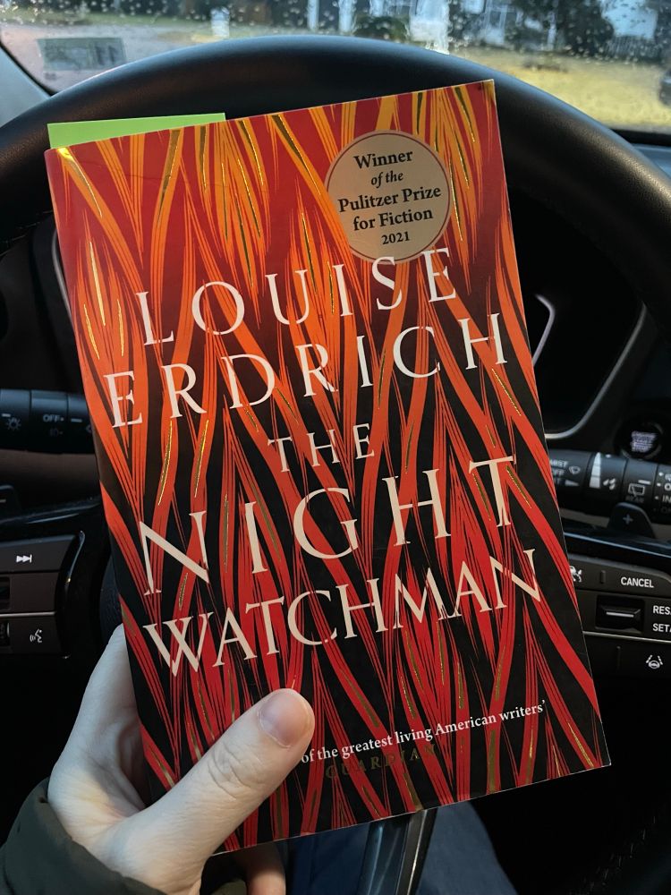 Hand holding a book in front of a car steering wheel 