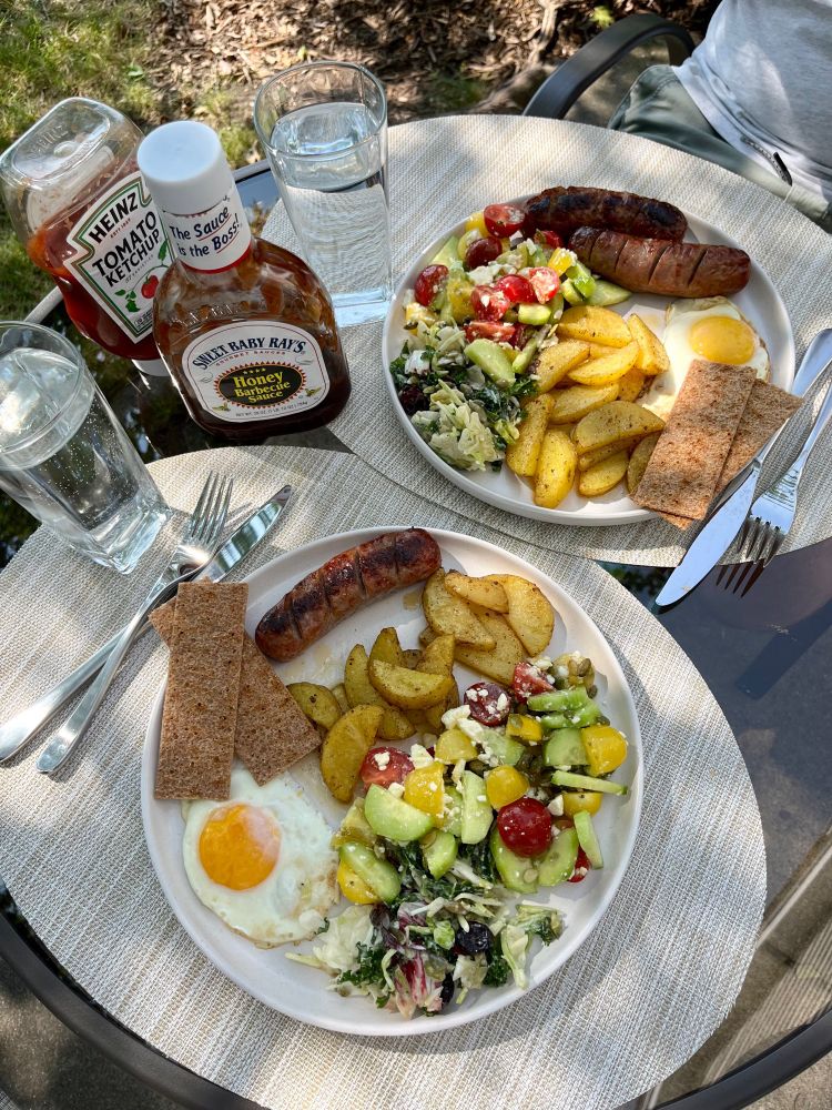 Фото сніданку. Дві тарілки з їжею: ковбаска, яйце, салати, хлібці.