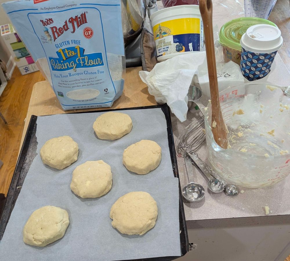 6 uncooked biscuits sitting on a baking pan next to a bowl of what previously had batter in it, a wooden spoon in the bowl, all sitting in front of a bag of gluten free Bob's Red Mill 1 to 1 baking flower.