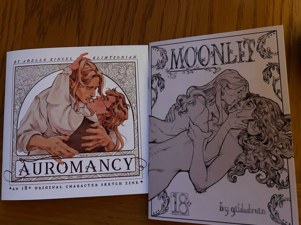 A photo of two zines. The one on the left is Auromancy by Klimtsonian. The one on the right is Moonlig by gildedruin. 