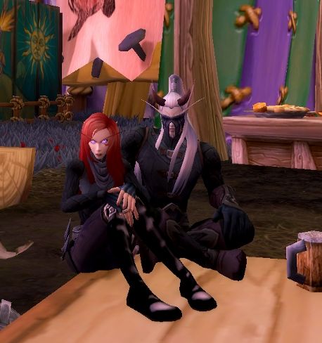 A screenshot from World of Warcraft depicting two blood elves sitting on a table. They are nearly matching in their attire, which is all black in both cases. One is a woman with bright red hair and the other is a demon hunter with short horns and long white hair.