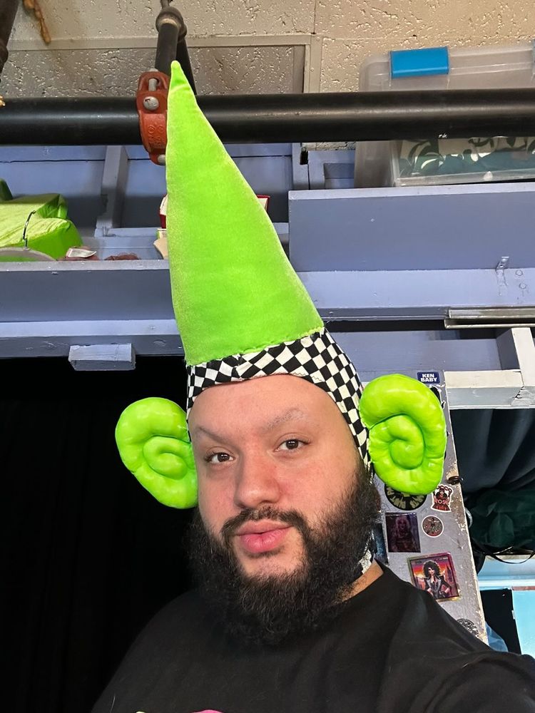 Just JP takes a selfie wearing no makeup and a head cap with two big stuffed green ears and an XL pointy green horn. Backstage at jacques.