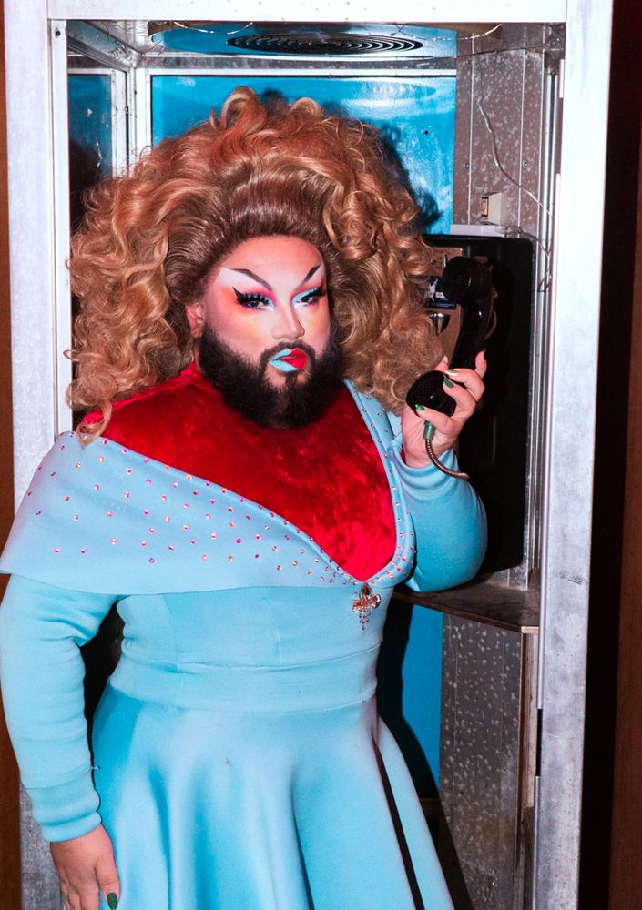 Just JP stuns in a Die With A Smile inspired outfit, wearing red under a blue dress and a dirty blonde large wig, with red and blue makeup to match. JP holds a phone to their face while staring down the camera