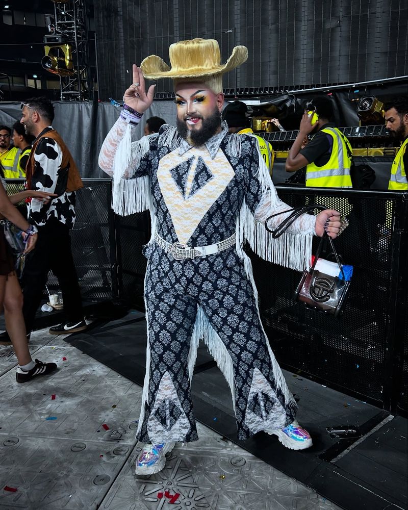Just JP poses in the pit after Beyonce's Cowboy Carter show, wearing a cowboy hat hair wig and a denim and white lace and fringe bodysuit