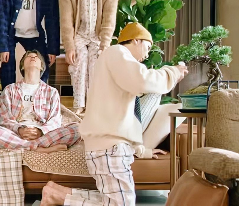 Namjoon, diligently spraying water on a bonsai, wearing a yellow beanie, a beige sweater and checkered pyjama pants