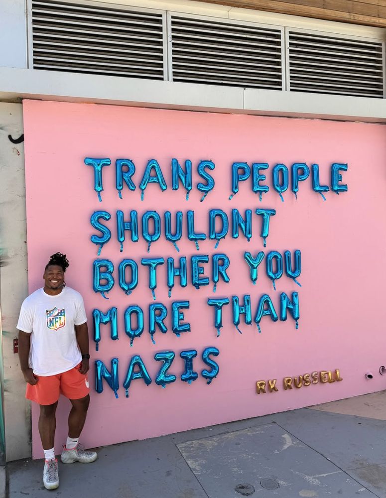 Ryan K. Russell standing in front of a pink wall with ballons that make up the statement saying: "Trans people shouldnt bother you more than Nazis"