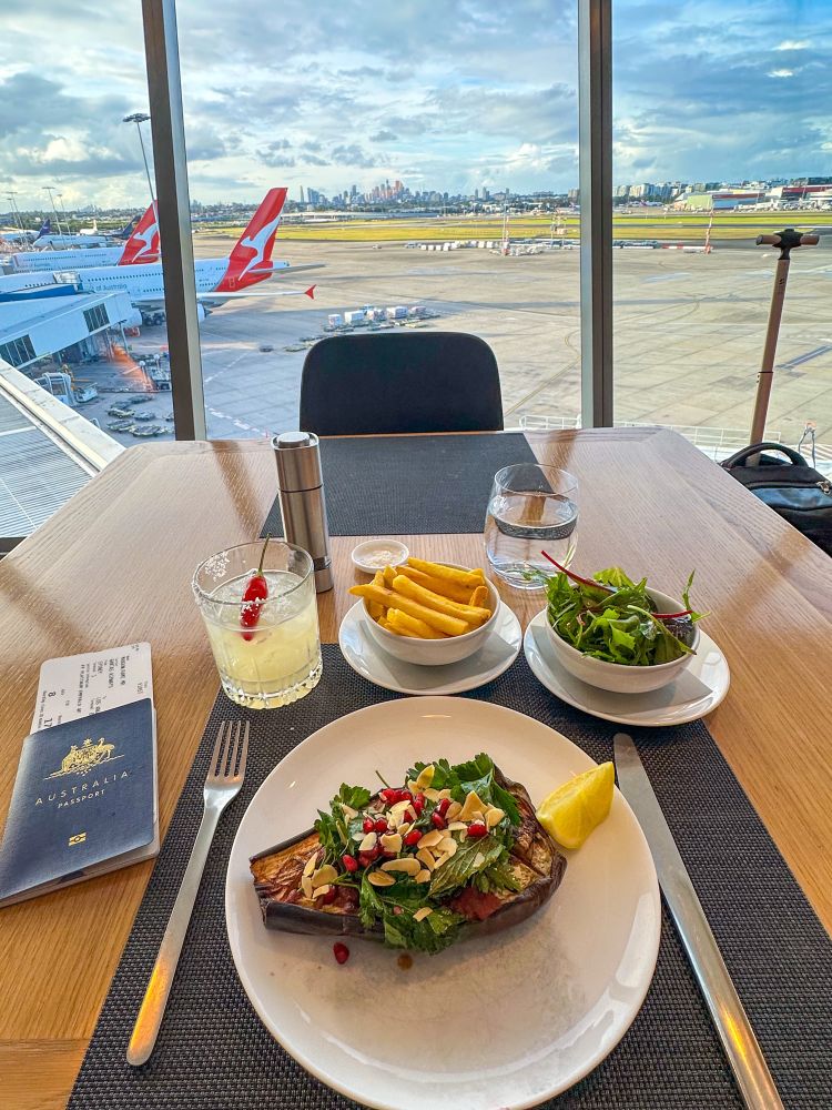 Several lunch dishes served at the airport lounge. There is also a cocktail. Outside the lounge and in the distance are several aircraft. 