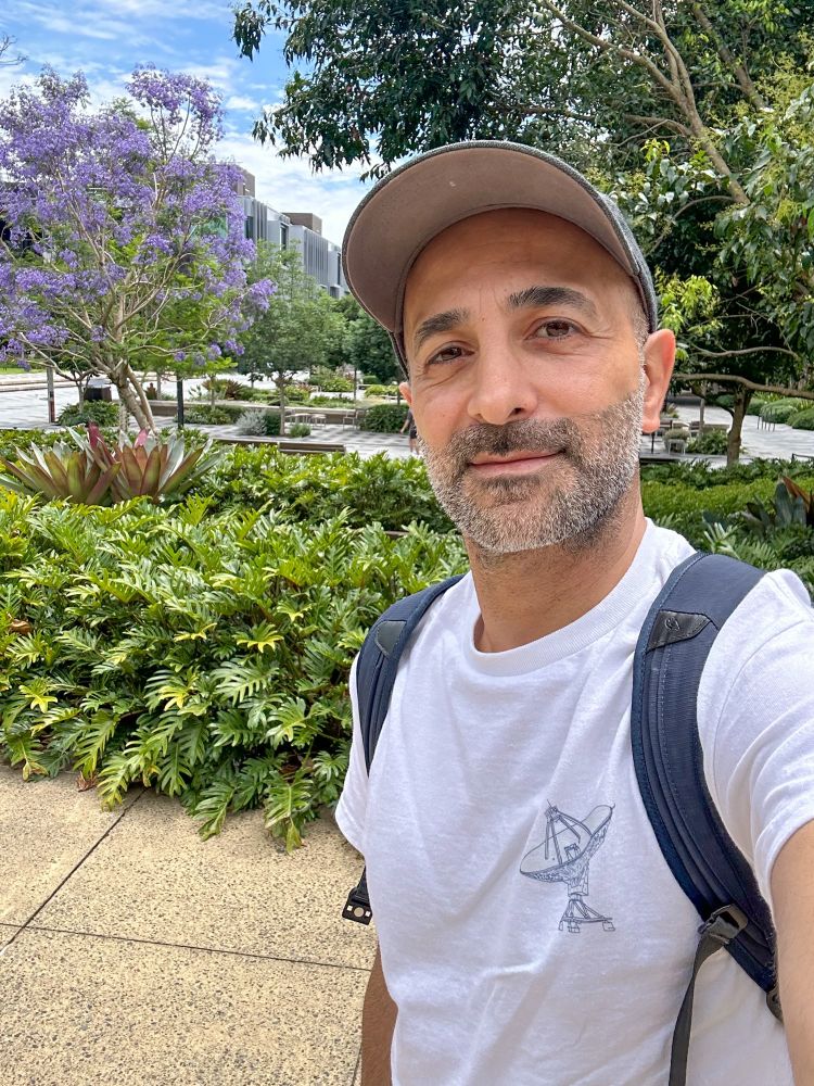 Selfie of me wearing a tshirt with a radio antennna on it and I am wearing a cap. Photo is taken in front of our campus garden and quad field. 