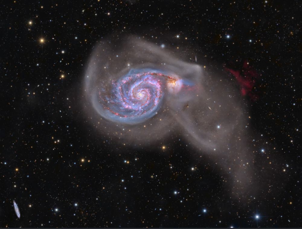 a large, spiral galaxy in the centre of the image, with wispy and diffuse nebulosity surrounding it and trailing off to the lower right. In the background are many stars of different colours, and a narrow cigar-shaped galaxy in the distance. 