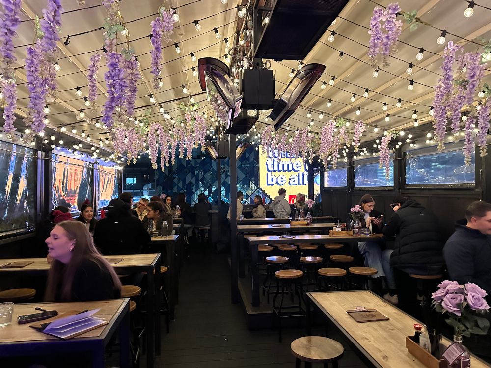 Looking out across a rooftop bar in Melbourne. The roof is covered and hanging are bunches of purple flowers and lights. There are people at tables eating and socialising. 