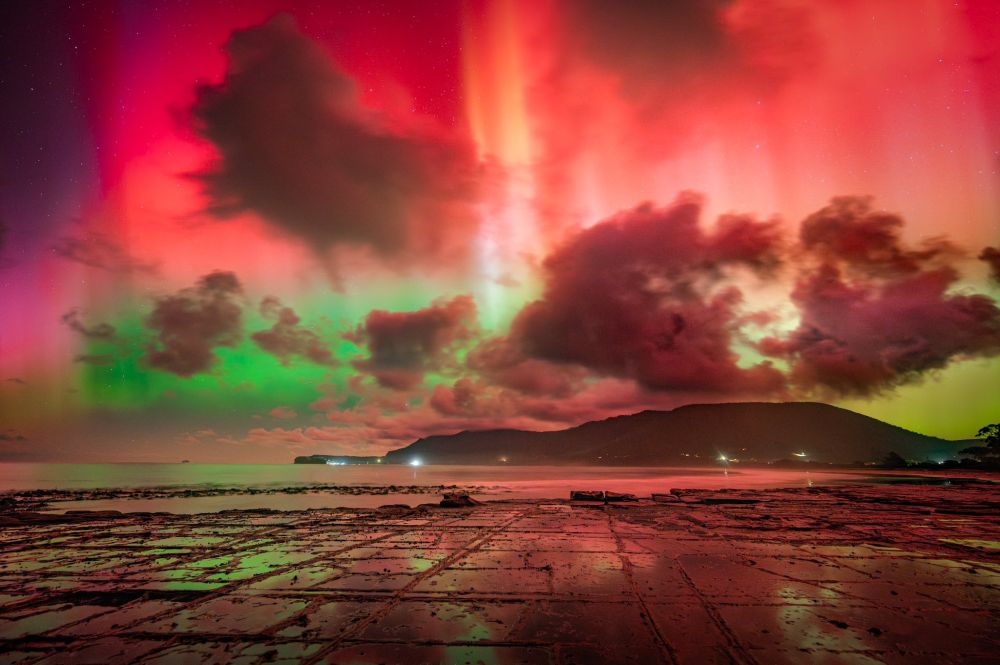 looking out over a natural flat rock shell towards a small hill. There are some clouds that are being silouetted against the bright red and green aurora