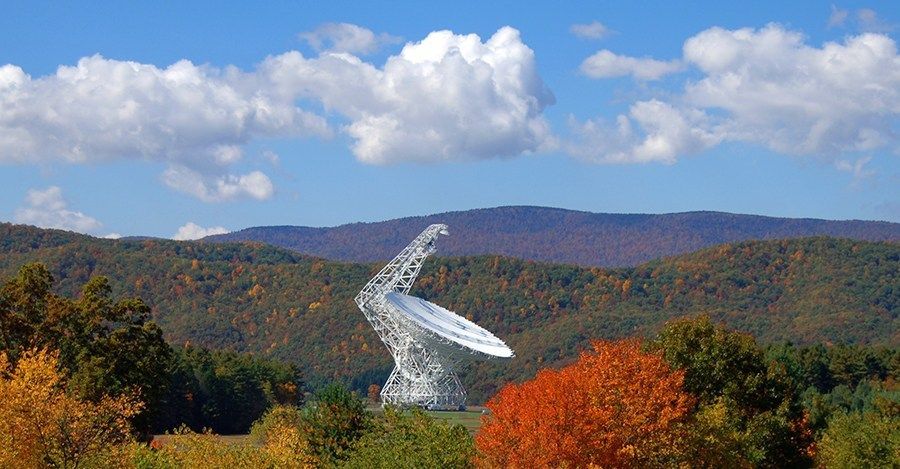 a large, white dish antenna telescope in the distance between the rolling hills. The trees in foreground and background are both golden amber as well as green.