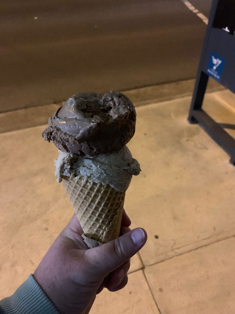 A hand holds an ice cream cone with two scoops of ice cream under a streetlight. The top scoop is choc malt, the second scoop is earl grey chocolate. 