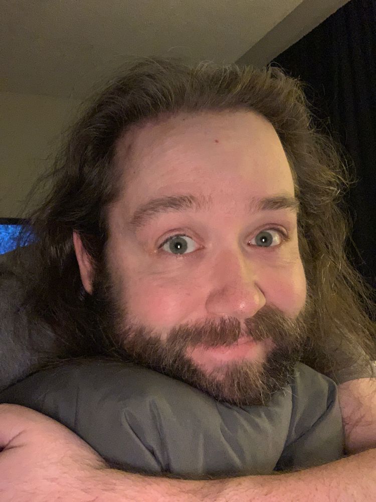 A selfie of Alec lying prone on his bed hugging a pillow under his chin. He makes a bear face at the camera.