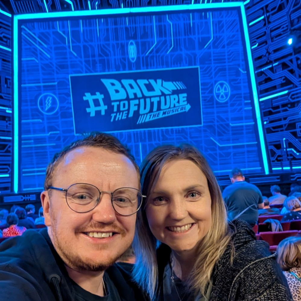 A woman and a man smiling in front of the blue starting screen for Back to the Future the Musical.
