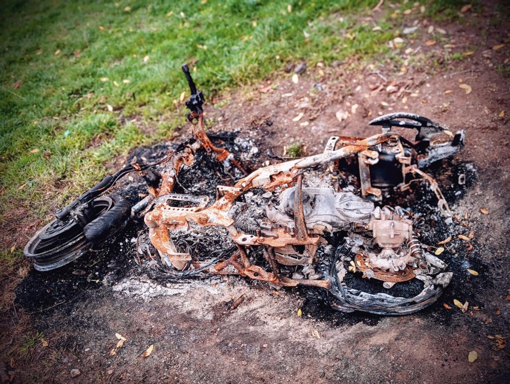 The burnt out, rusted shell of a motorbike lying in dirt, surrounded by grass and leaves.
