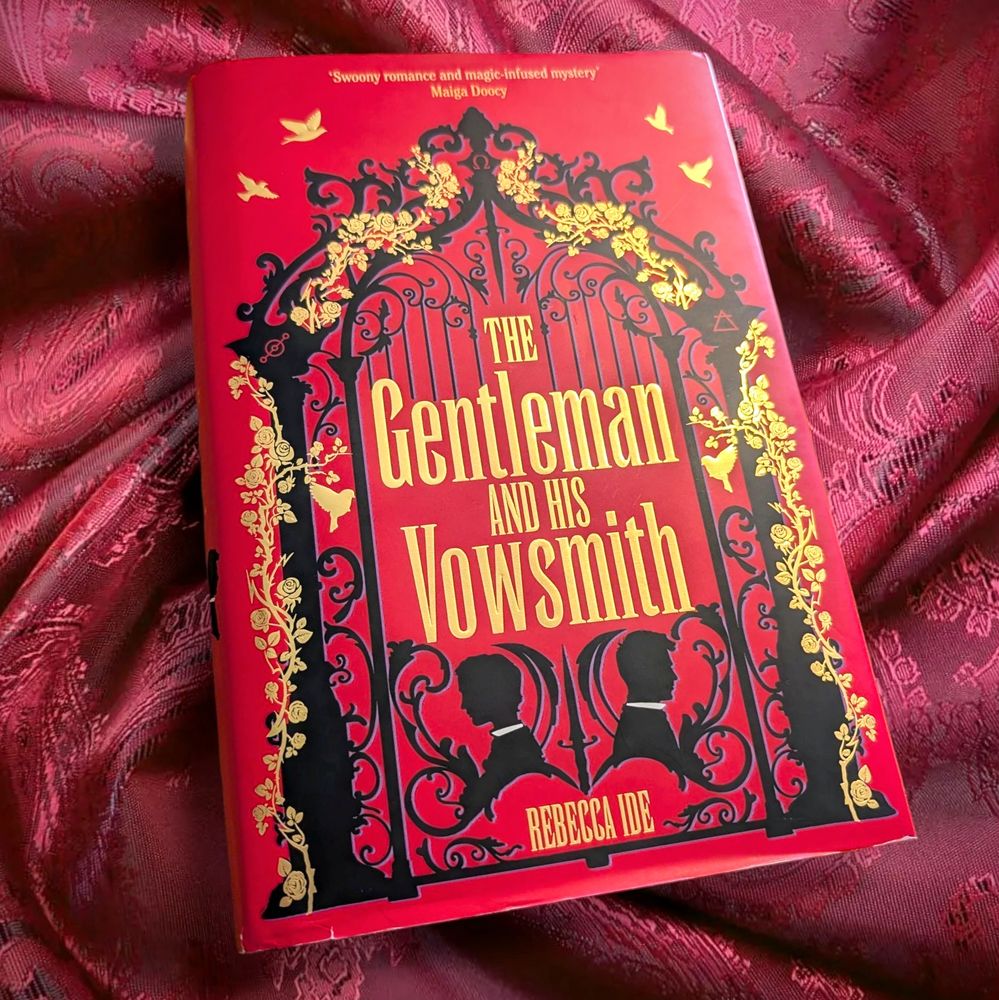 The cover of The Gentleman and His Vowsmith by Rebecca Ide against a red fabric background. The cover is bright red with black and gold detailing.