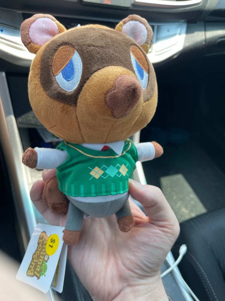 A Tom Nook plush toy being held in a hand. The tag is still attached and a “$1” sticker is attached to it.