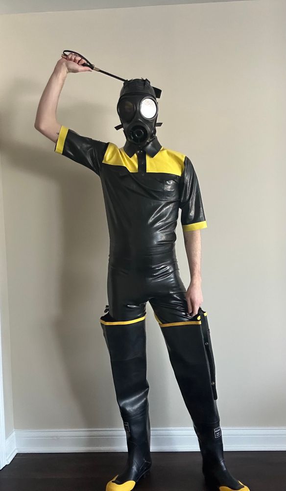 latex drone standing in black and yellow waders and rubber. a riding crop is held above their gas mask covered head. 
