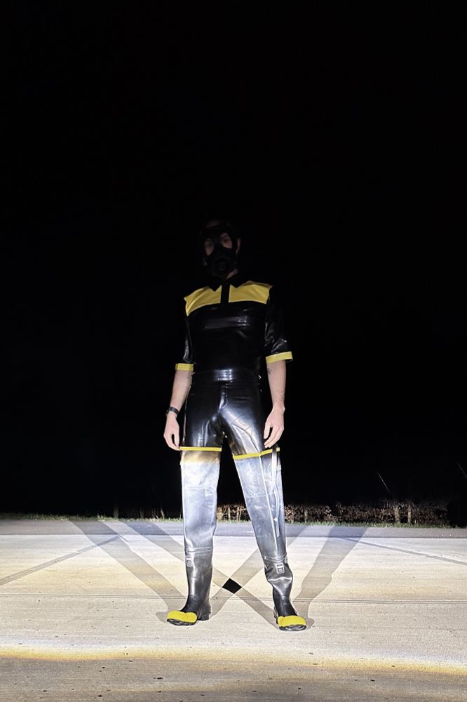 rubber drone in black rubber gear with yellow accents standing, illuminated by car headlights. 
