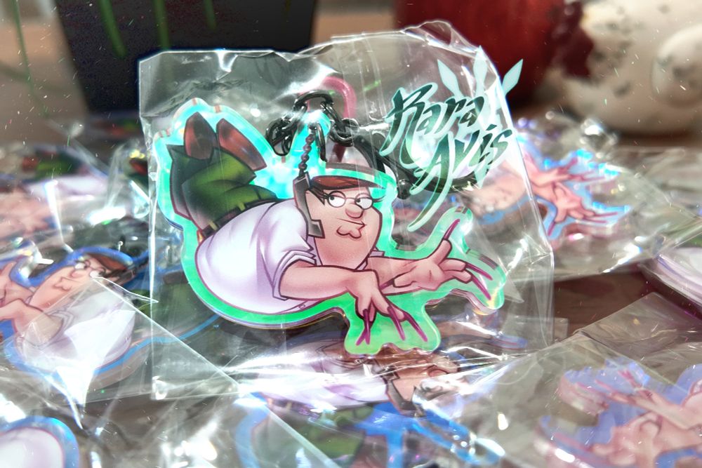 The previous illustration, but in keychain form. A gunmetal gray lobster hook clasp, circle split ring, and chain hang from the top of the phone cord, and the keychain is outlined by a rainbow cyan-green shimmer. The keychain is in a bag, and it stands on top of a pile of other keychains of the same type. The image is lightly edited for clarity. The watermark on the photo says "Rara Avis."