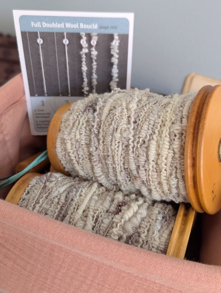 Close up photo of a pink fabric basket. Inside are two very full bobbins of boucle yarn and an instruction card on how to make double wool bouclé. The yarn of mostly off white wool with pops if burgundy silk. 