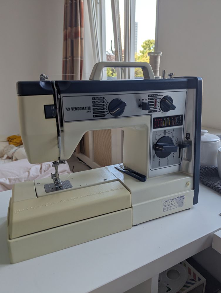 Shot of a retro looking sewing machine in blue and white colors. It's sat on a white table top and dominates the picture. 