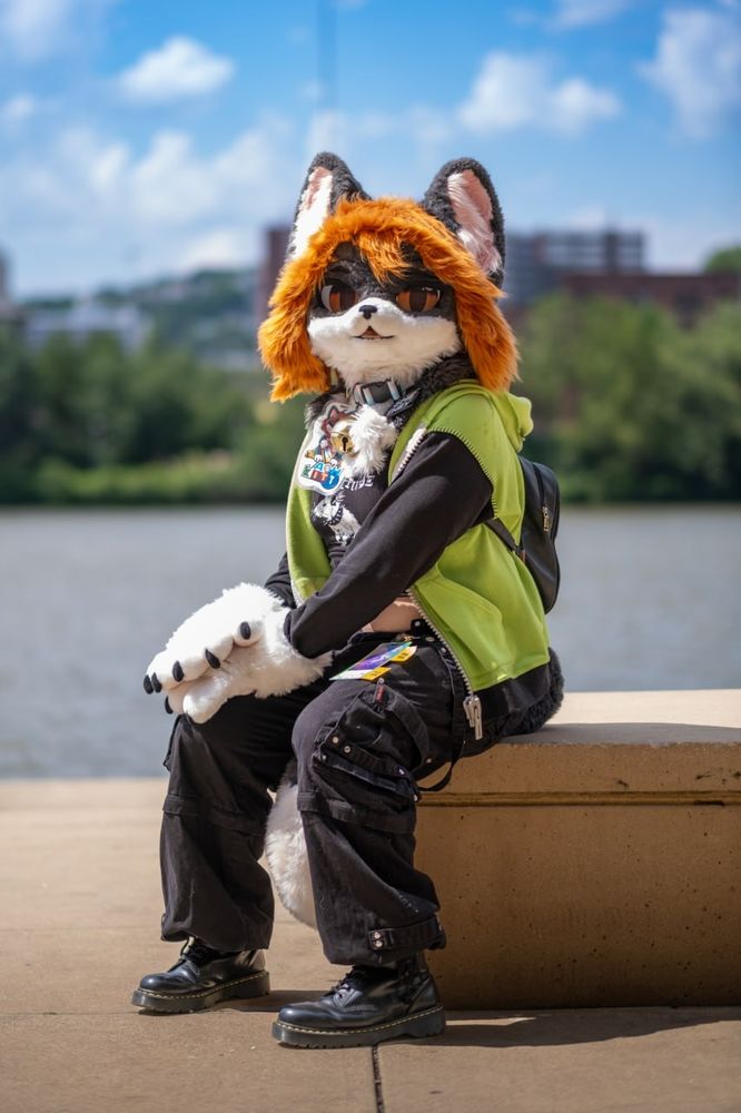 Me in my tuxedo cat fursuit, with a Gir hoodie, crop top, black Tripp pants, and Doc Martens sitting on a concrete bench-like structure on the waterfront underneath the David Lawrence Convention Center