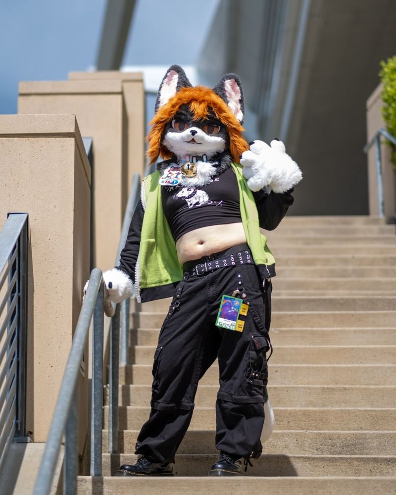 Me in my tuxedo cat fursuit, with a Gir hoodie, crop top, black Tripp pants, and Doc Martens posing on a staircase on the waterfront underneath the David Lawrence Convention Center