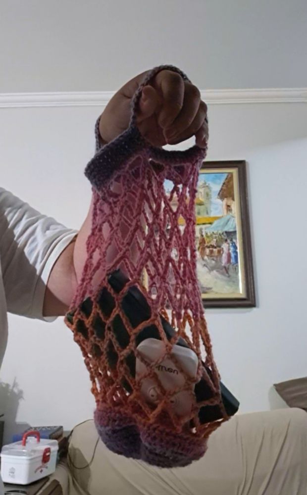 Bolsa formada, ela é pequena, tem as cores laranja, rosa e roxa, alças curtas e é vazada, em formato de losangos. Além disso segura um controle remoto e uma caderneta.