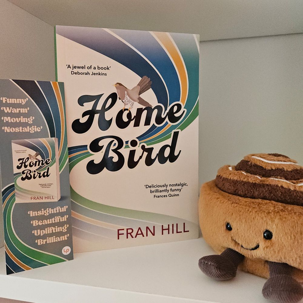 Copy of the book Home Bird standing alongside a cinnamon bun jellycat and a themed bookmark on a white bookshelf