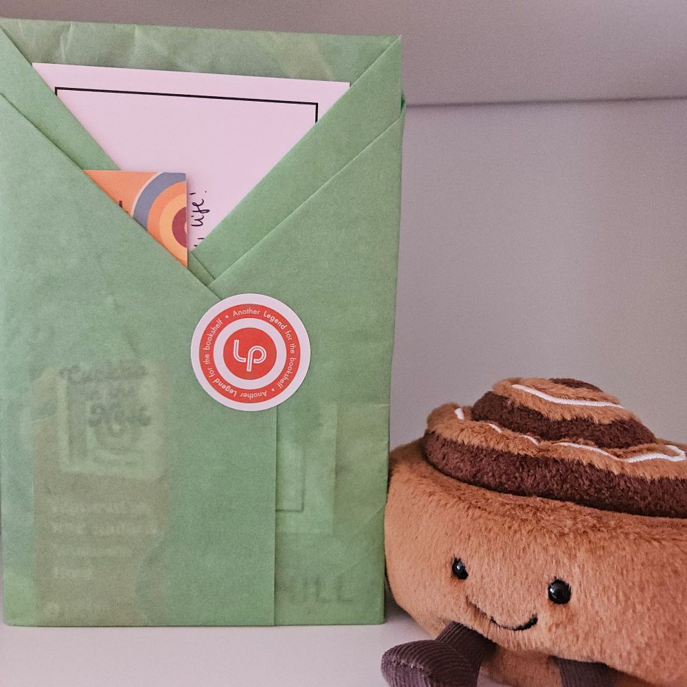 Book wrapped in pale green tissue stading alongside a cinnamon bun jellycat on a white bookshelf