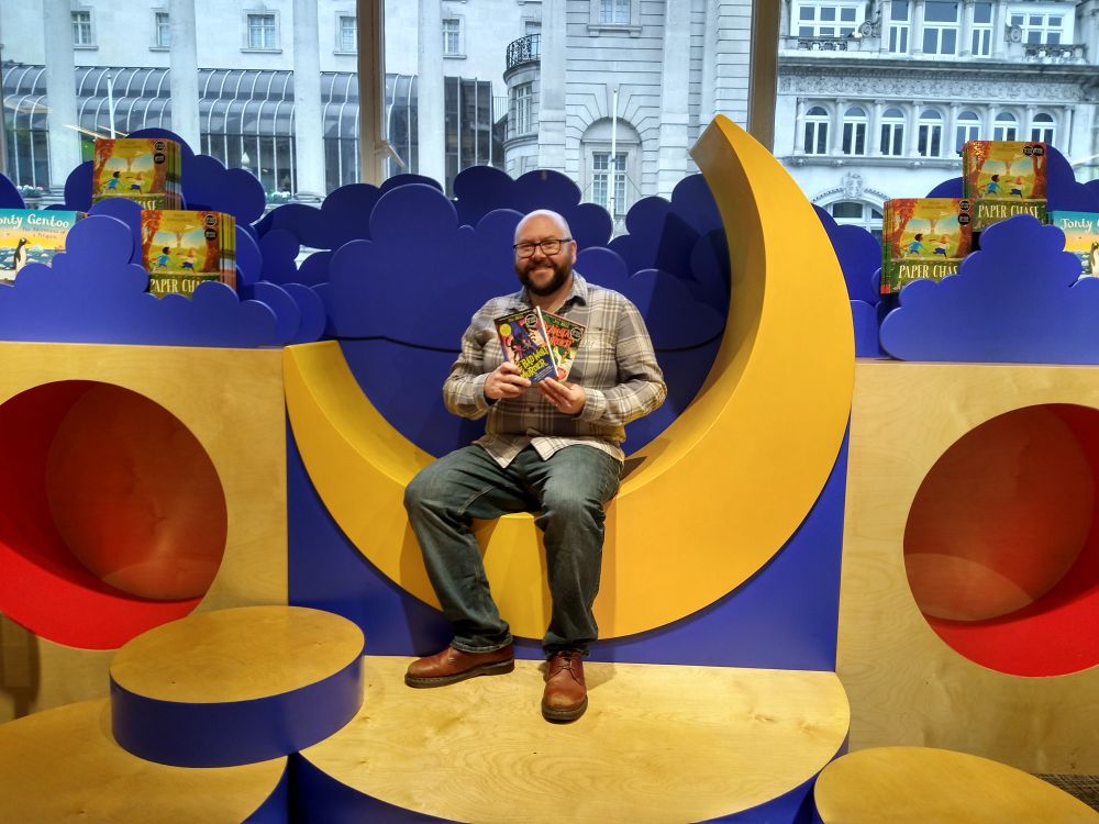 Author P.G. Bell sitting in a moon-shaped seat in Waterstones Piccadilly. He's holding copies of his books, The Big Bad Wolf Murder and The Beanstalk Murder.
