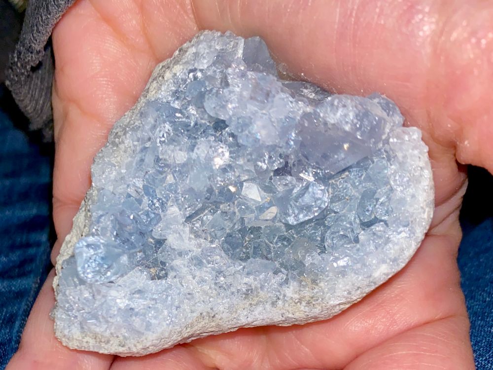 A light blue raw crystal geode piece in the rough shape of a heart sparkles in the palm of a hand. 