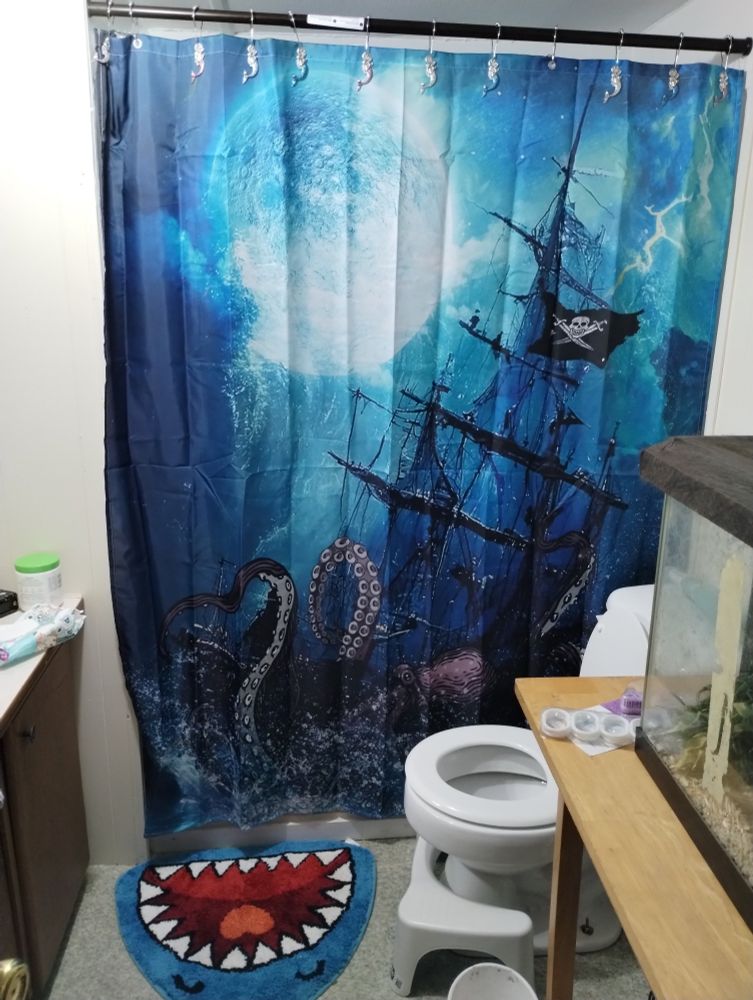 A bathroom with a shark bath mat and a shower curtain with a pirate ship being drug down by a kraken. The curtain holders are little mermaids