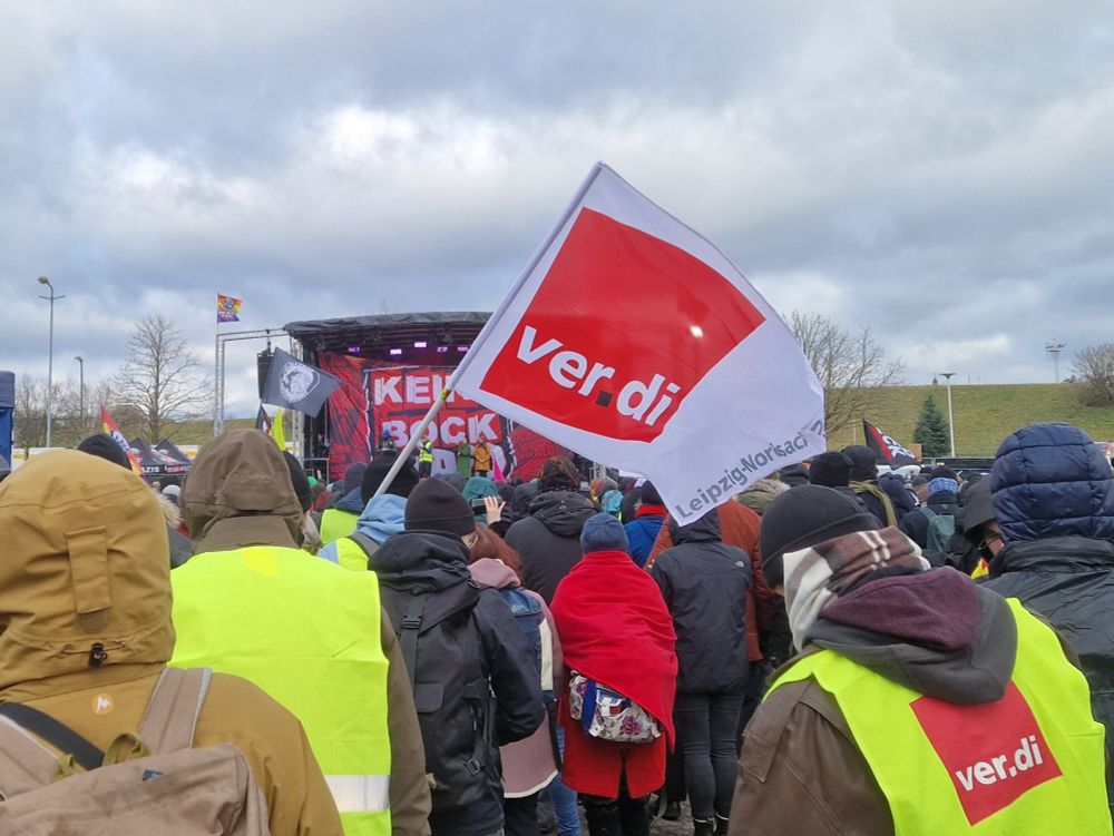Auf dem Bild sind Demoteilnehmer von hinten und eine verdi-Fahne zu sehen. Im Hintergrund die Bühne.
