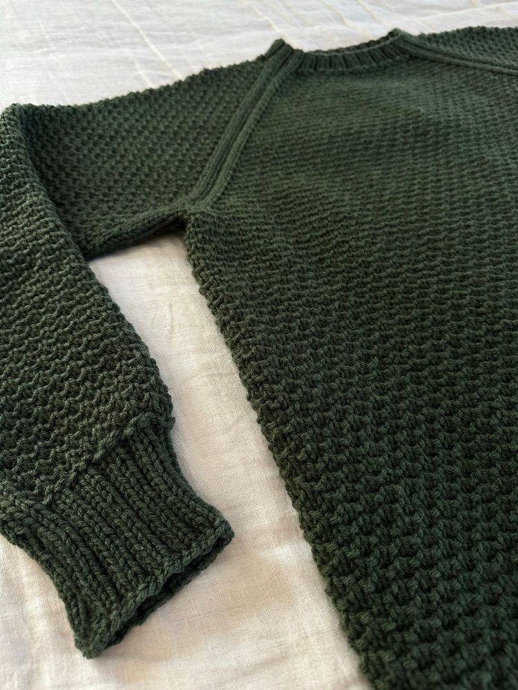 A green, textured hand knit sweater laying flat on a linen duvet. 